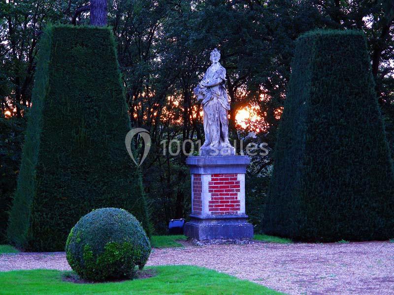 Statue en pierre sur un socle en brique, entourée de haies taillées et d'arbres au coucher du soleil.