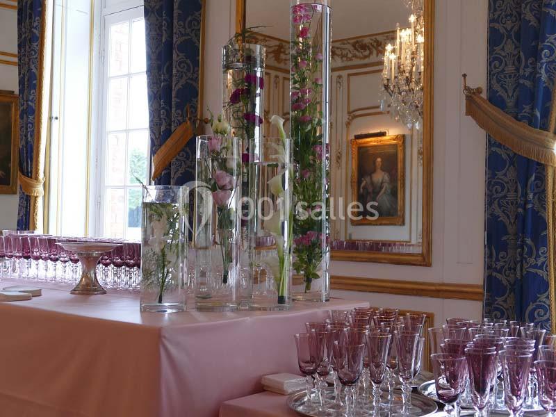 Table ornée de verres violets et de vases avec des fleurs, dans une salle élégante avec miroirs et lustres.