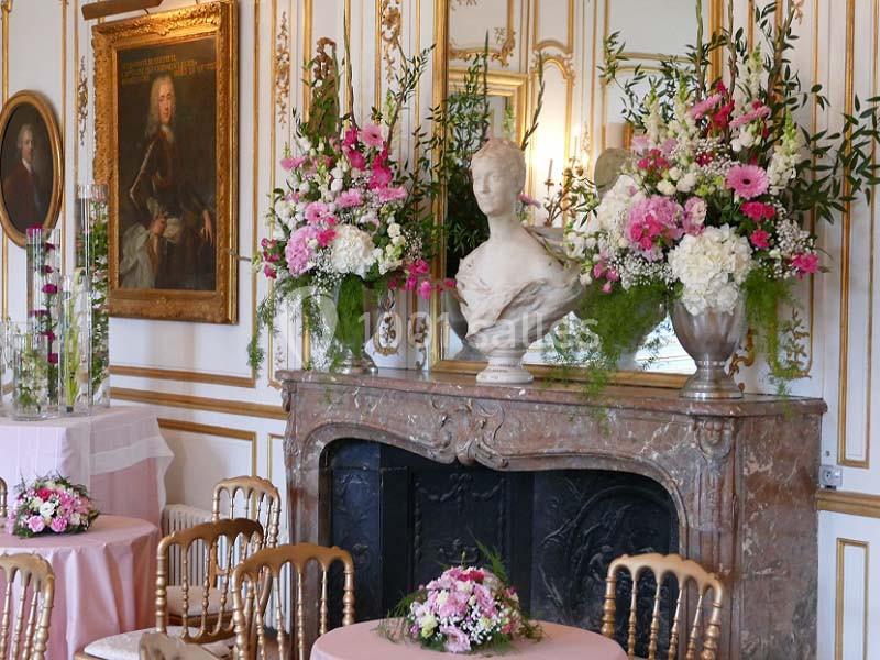Salle élégante avec cheminée en marbre, bustes, tableaux anciens et décorations florales roses et blanches.
