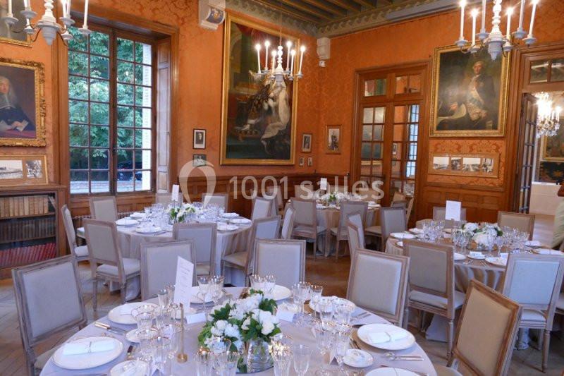 Salle élégante avec tables rondes dressées pour un repas, décorée de tableaux, chandeliers et grandes fenêtres.
