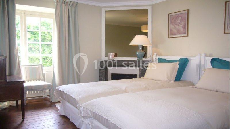 Deux lits simples avec linge blanc dans une chambre lumineuse, décorée de rideaux clairs et d'une lampe sur une cheminée.