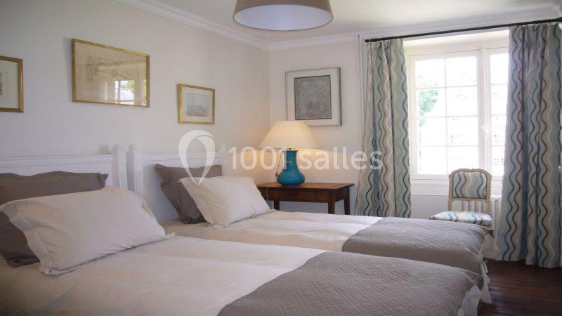 Chambre lumineuse avec deux lits simples, une lampe bleue sur une table en bois et des rideaux à motifs près d'une fenêtre.