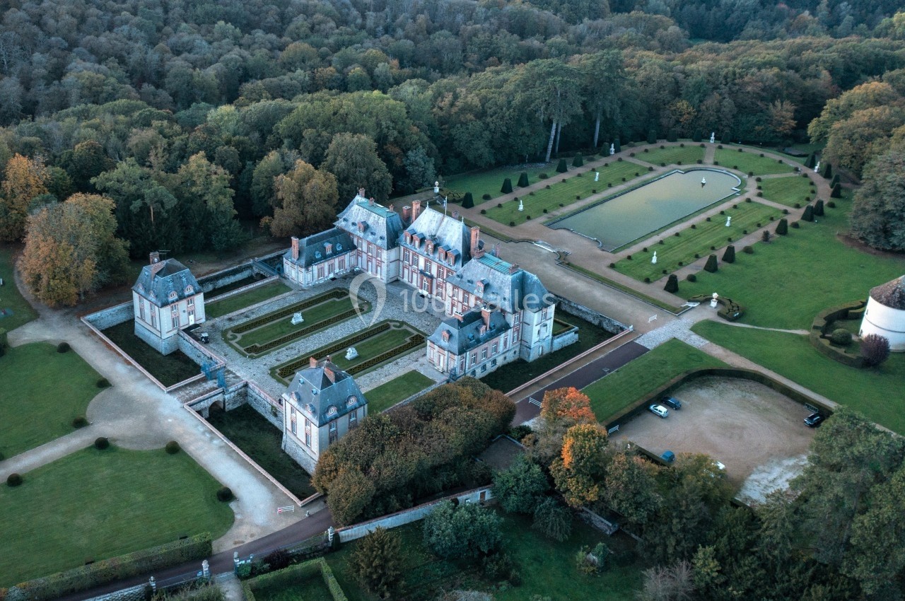 Vue aérienne d'un château entouré de jardins symétriques, d'un étang et d'une forêt en arrière-plan.