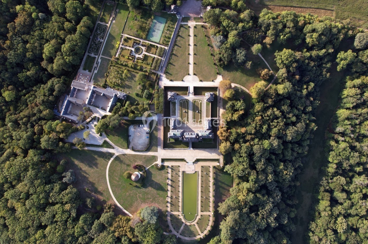 Vue aérienne d'un château entouré de jardins symétriques, bassins et forêt dense.