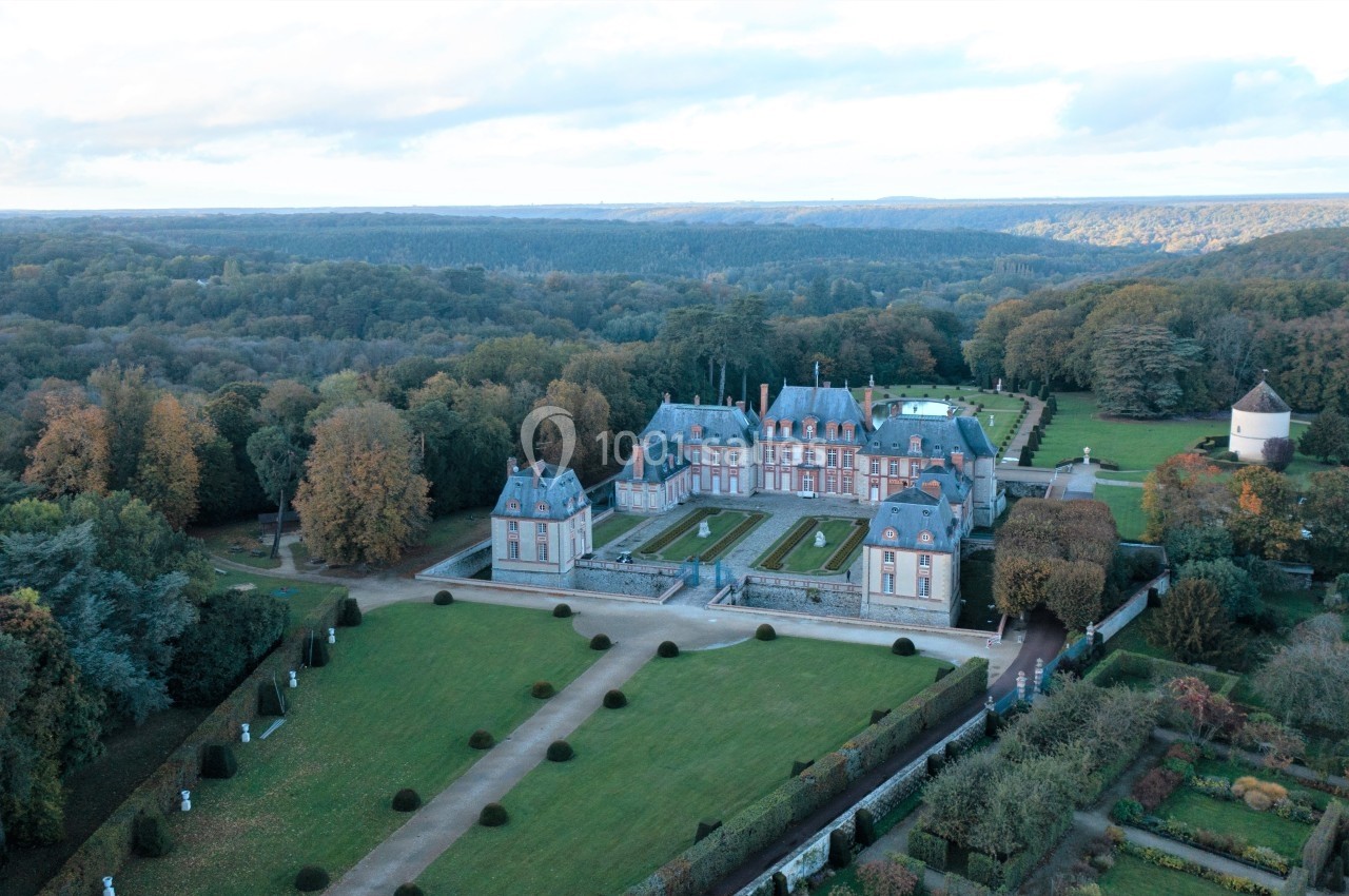 Vue aérienne d'un château entouré de jardins symétriques, d'arbres et d'une campagne boisée.