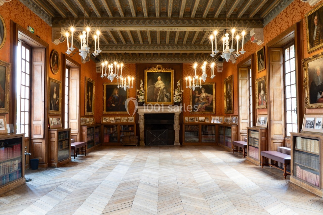 Salle ornée de portraits et de chandeliers, avec parquet en bois, bibliothèque et cheminée centrale.