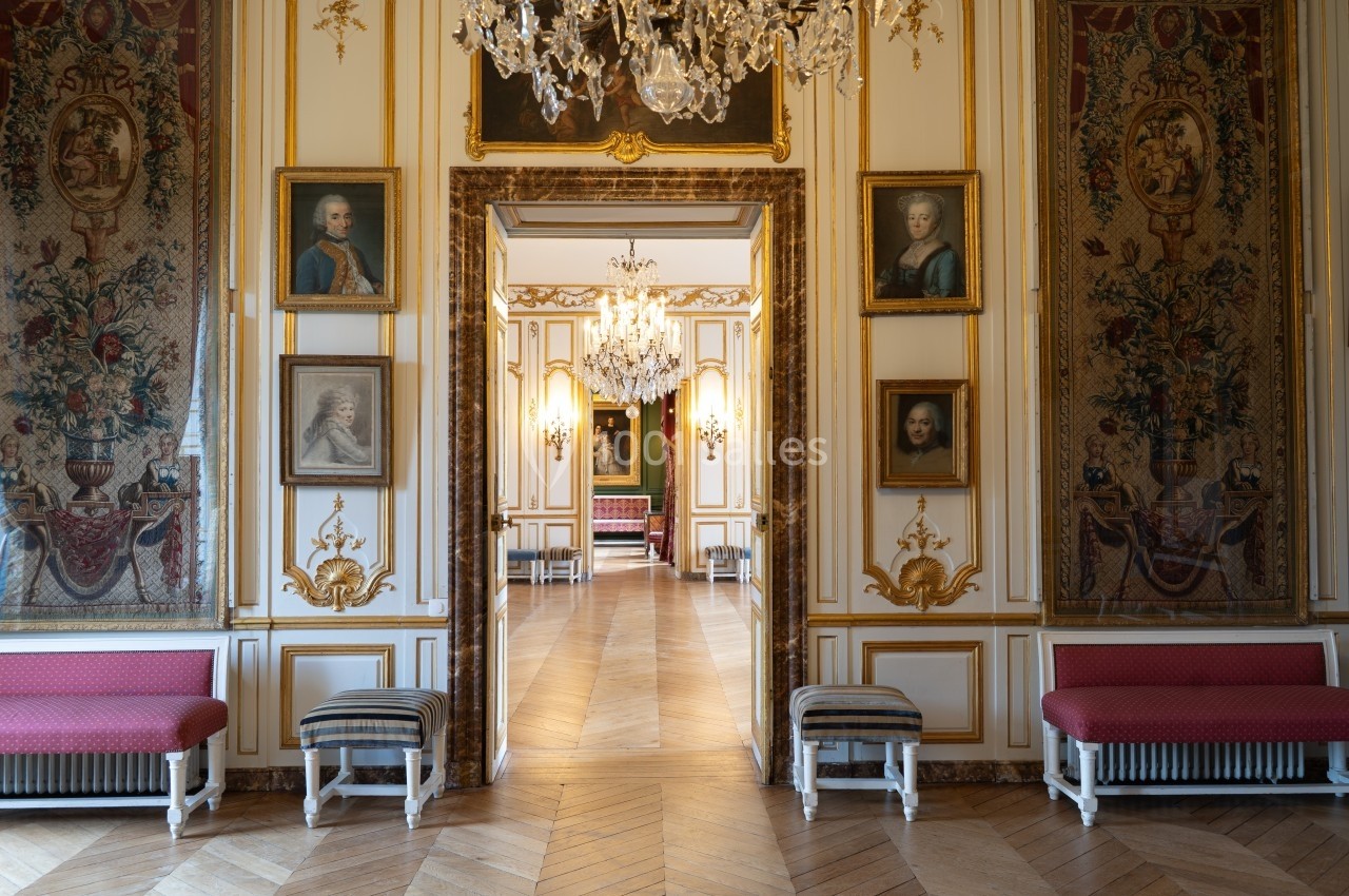 Intérieur d'un palais avec parquet en bois, lustres en cristal, portraits encadrés et tapisseries ornées.