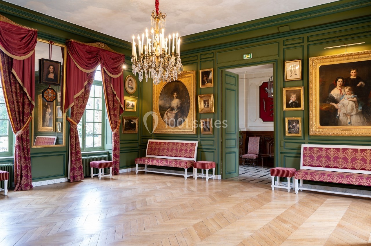 Salle ornée de peintures classiques, avec murs verts, rideaux rouges, parquet en bois et un lustre central.