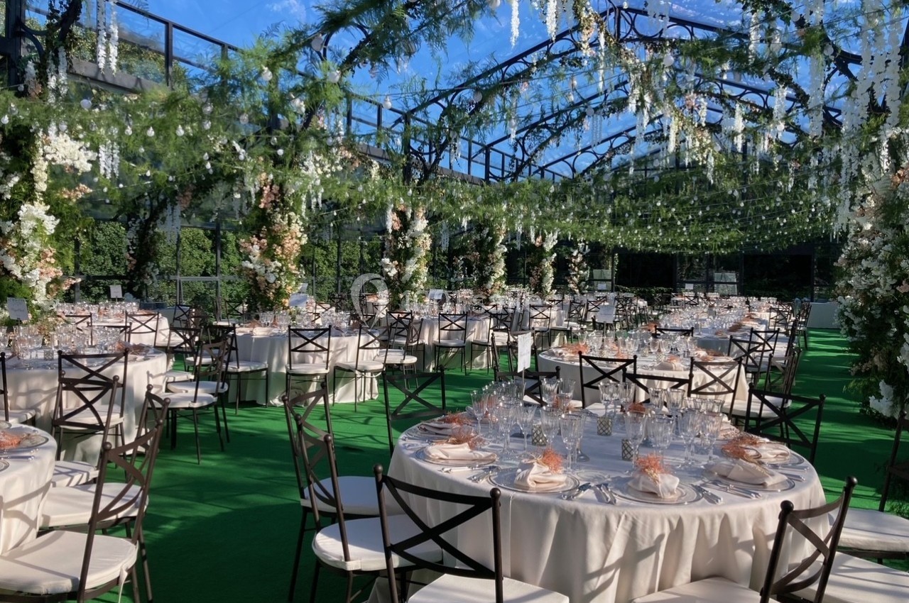 Salle de réception en plein air décorée de guirlandes florales, avec des tables rondes dressées sur une pelouse verte.