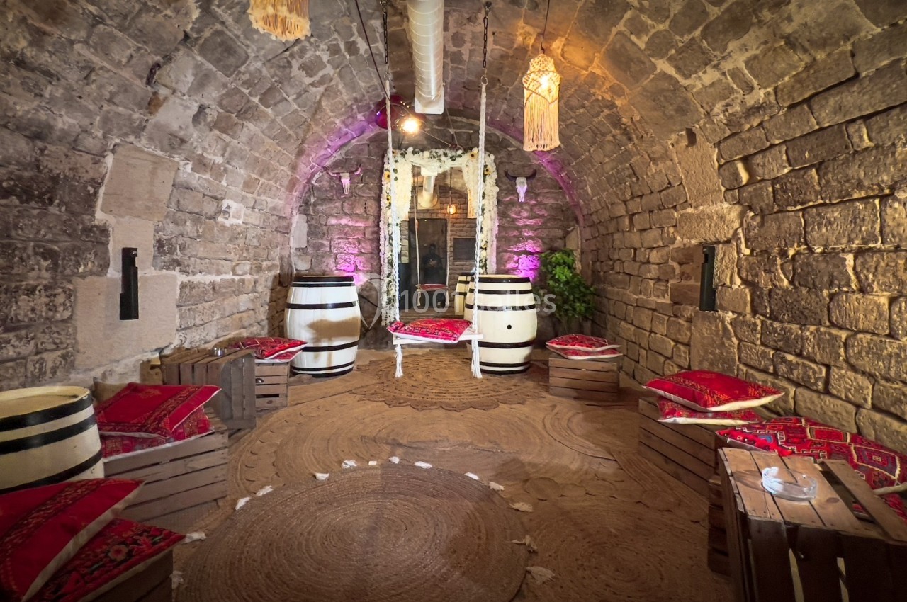 Salle voûtée en pierre avec des tonneaux, coussins colorés et éclairage tamisé, décorée dans un style rustique.