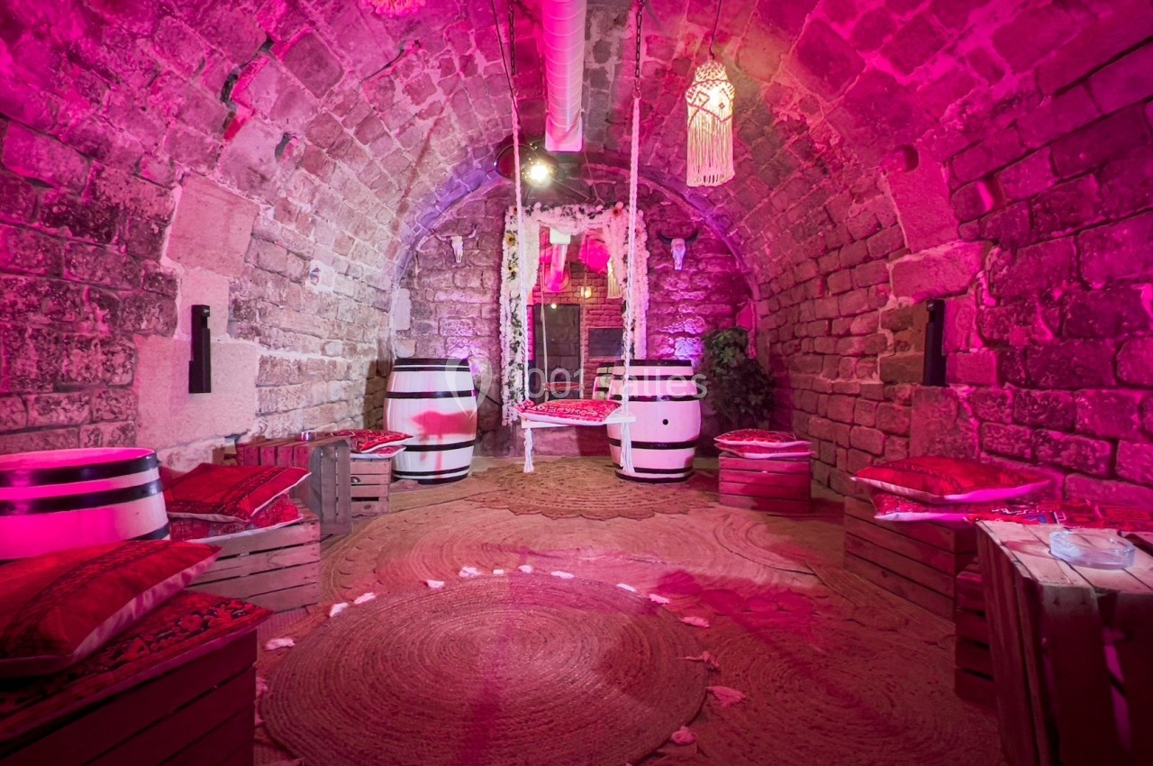 Salle voûtée en pierre avec éclairage rose, décorée de coussins, tonneaux et hamacs suspendus.