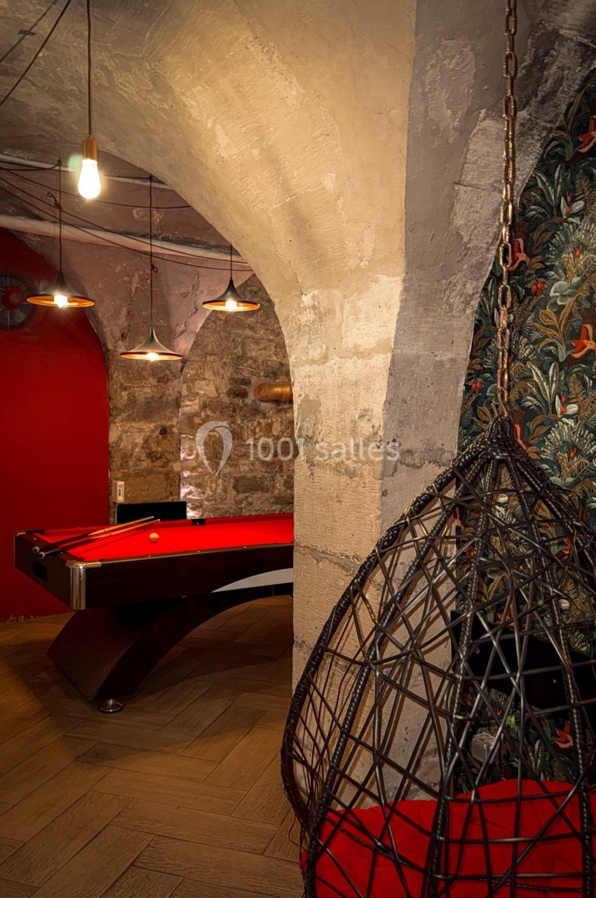 Salle voûtée avec un billard rouge, un fauteuil suspendu en osier et des murs en pierre apparente.