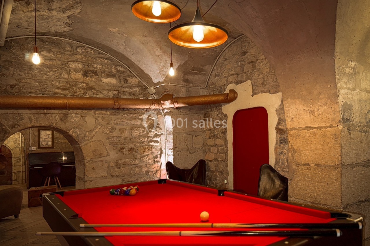 Table de billard rouge dans une pièce voûtée en pierre, éclairée par des suspensions lumineuses.