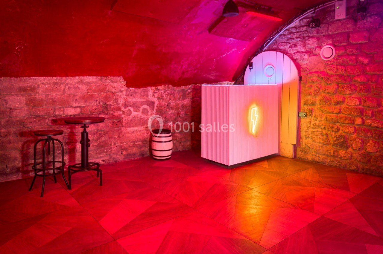 Salle voûtée en briques avec éclairage rouge, sol en bois, table haute, tabourets et comptoir lumineux avec un néon.