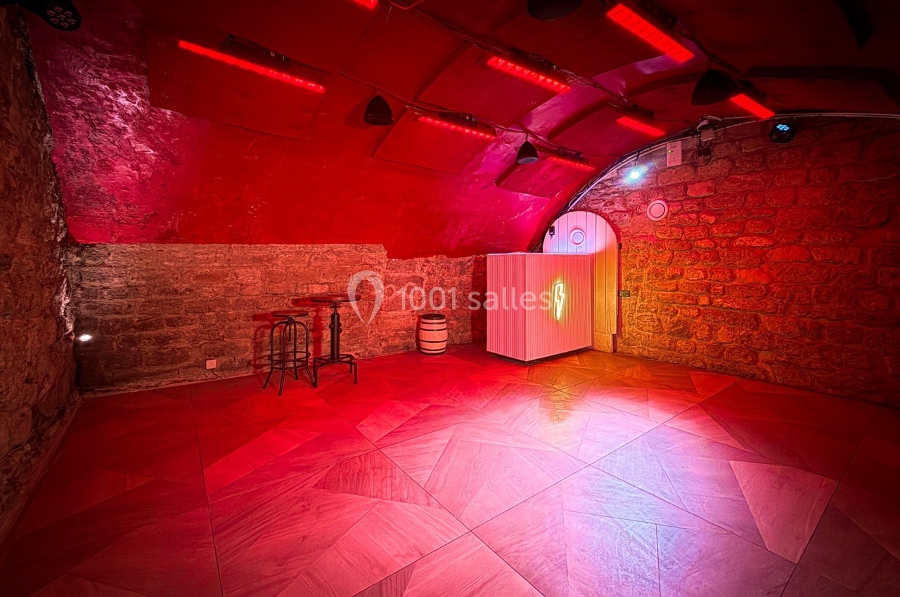 Salle voûtée en pierre éclairée par des lumières rouges, avec un bar, des tabourets et un tonneau en décoration.