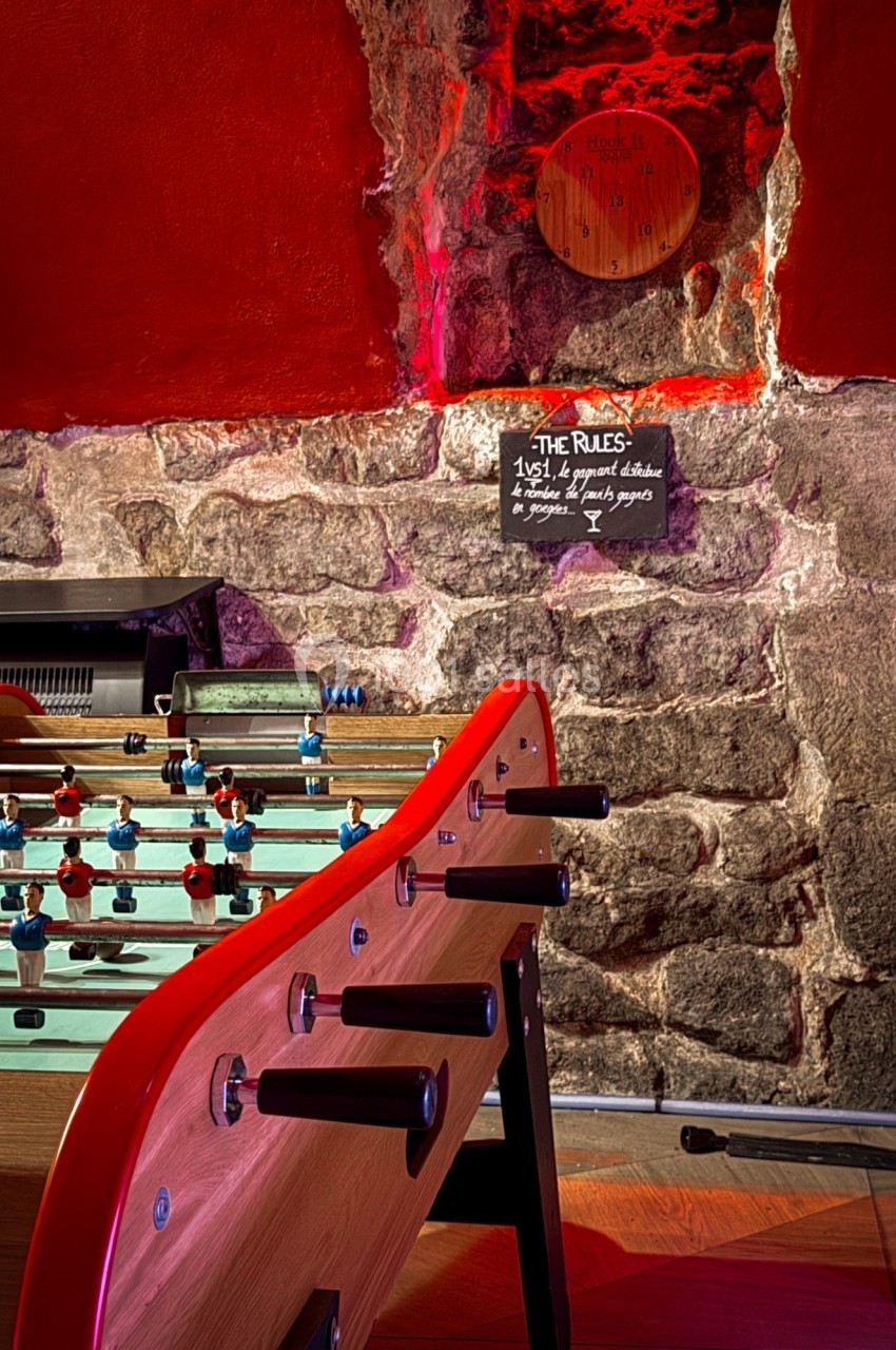 Baby-foot en bois dans une pièce aux murs en pierre, éclairée par une lumière rouge, avec un tableau de règles au mur.