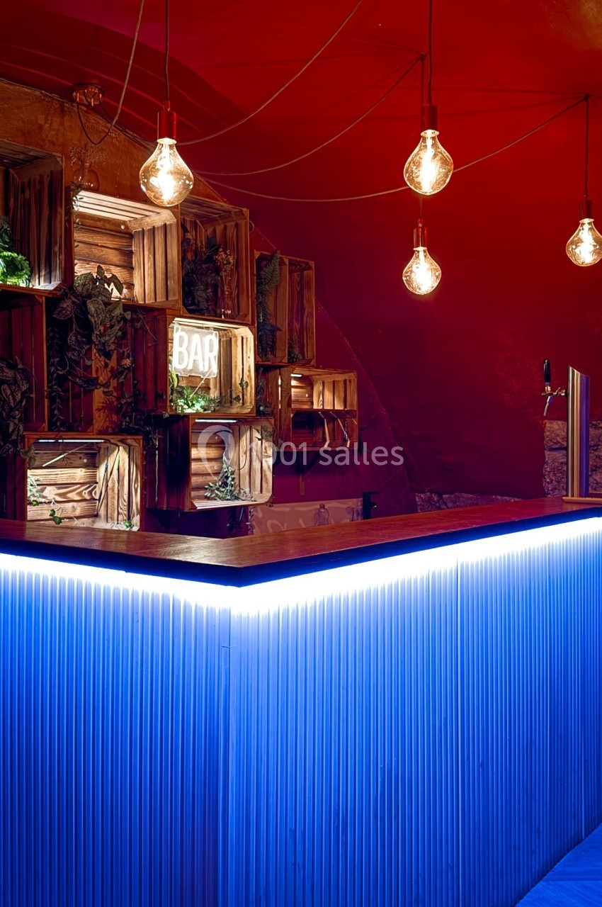 Comptoir de bar éclairé par une lumière bleue, avec des ampoules suspendues et des caisses en bois décoratives au mur.