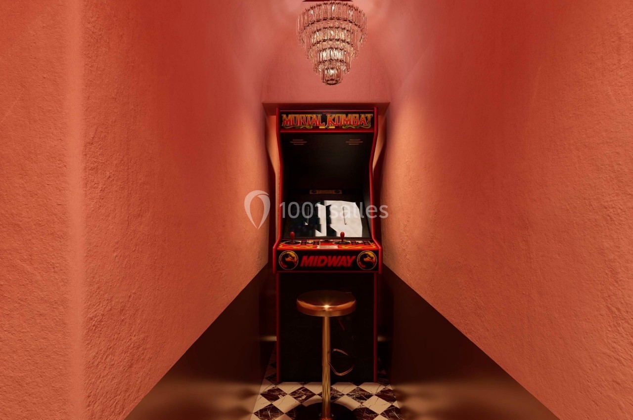 Borne d'arcade rétro Mortal Kombat dans un couloir étroit aux murs roses, éclairée par un lustre.