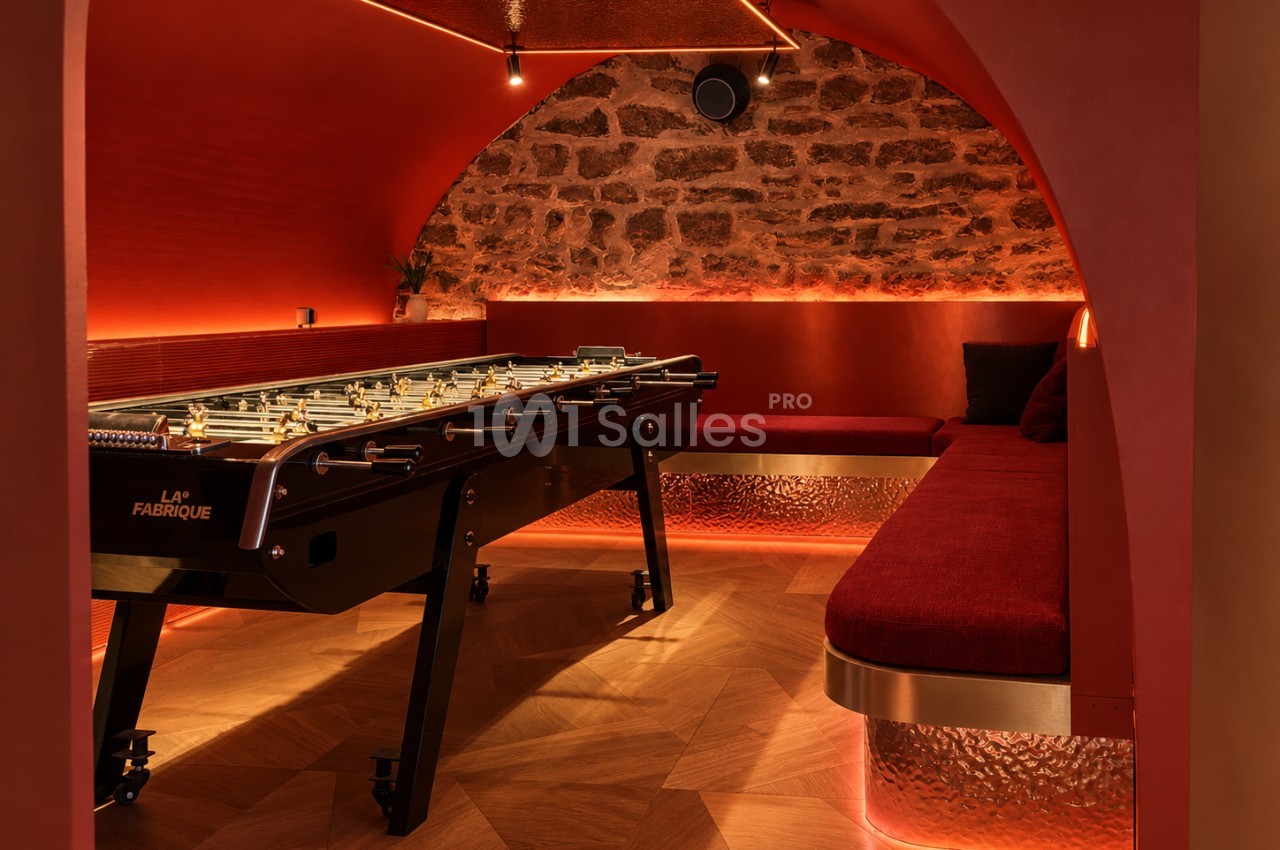 Salon voûté aux murs rouges éclairés en contre-jour, avec un baby-foot au premier plan et une banquette rouge le long d'un mur en pierre apparente.