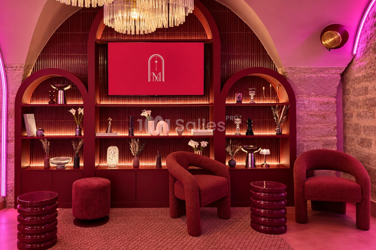 Salon aux tons bordeaux et roses éclairé au néon, avec deux fauteuils bouclés, des poufs et un mur de niches arrondies garnies de vases et d'objets, sous un lustre à pampilles.