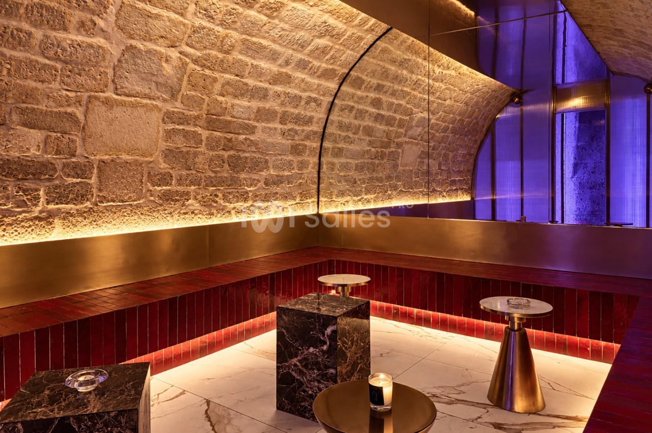Salle voûtée aux murs en pierre apparente éclairés en contre-jour, banquettes recouvertes de carreaux rouges, tables basses en marbre et en laiton sur un sol en marbre clair.