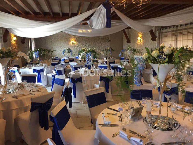 Salle de réception décorée avec des tables rondes, nappes blanches, chaises ornées de nœuds bleus et compositions florales.