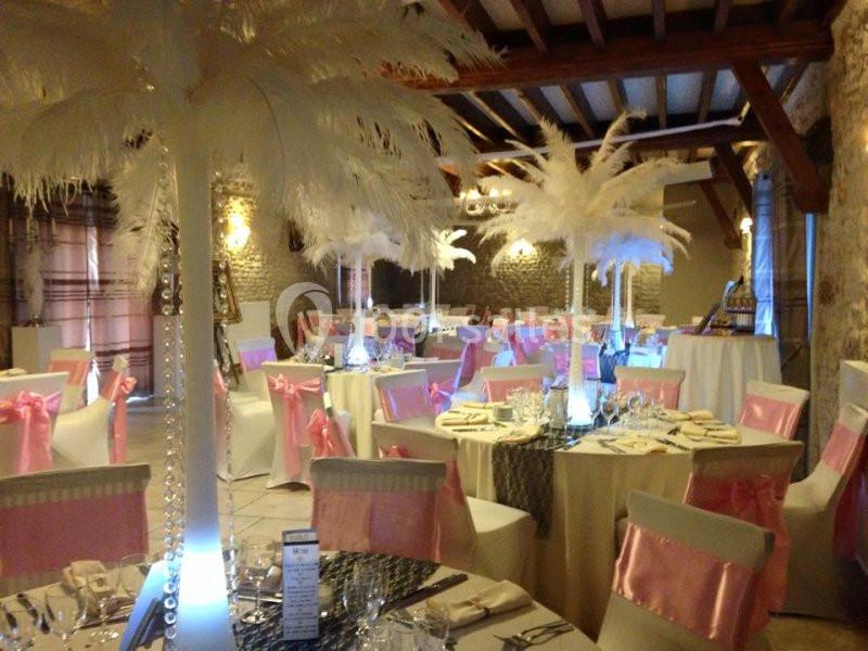 Salle de réception décorée avec des tables dressées, des chaises ornées de nœuds roses et des centres de table à plumes…