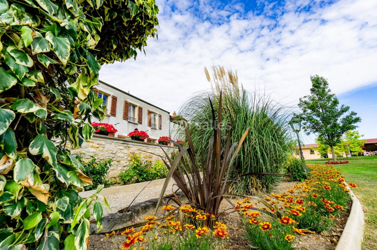 Jardin fleuri avec des plantes et des fleurs colorées devant une maison en pierre sous un ciel partiellement nuageux.