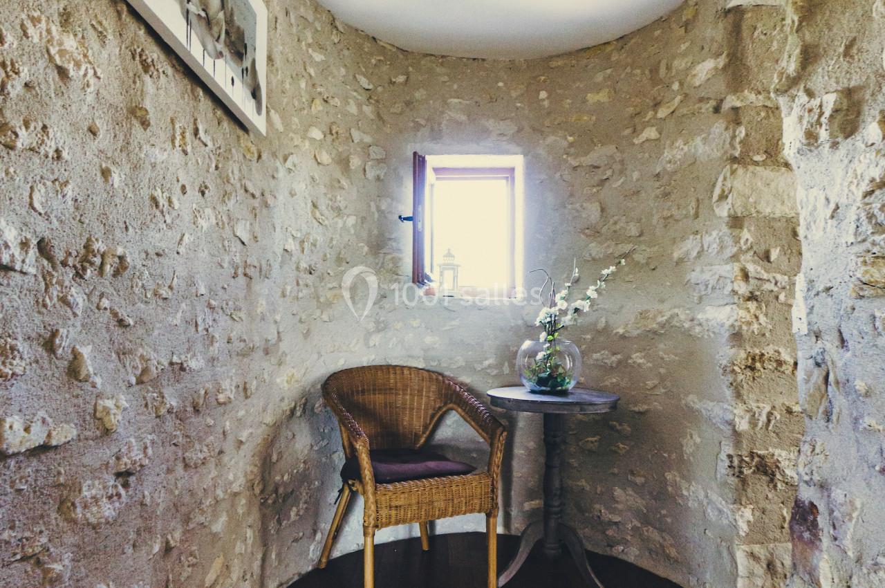 Coin d'une pièce en pierre avec une chaise en osier, une petite table ronde, un vase de fleurs et une fenêtre éclairant l…