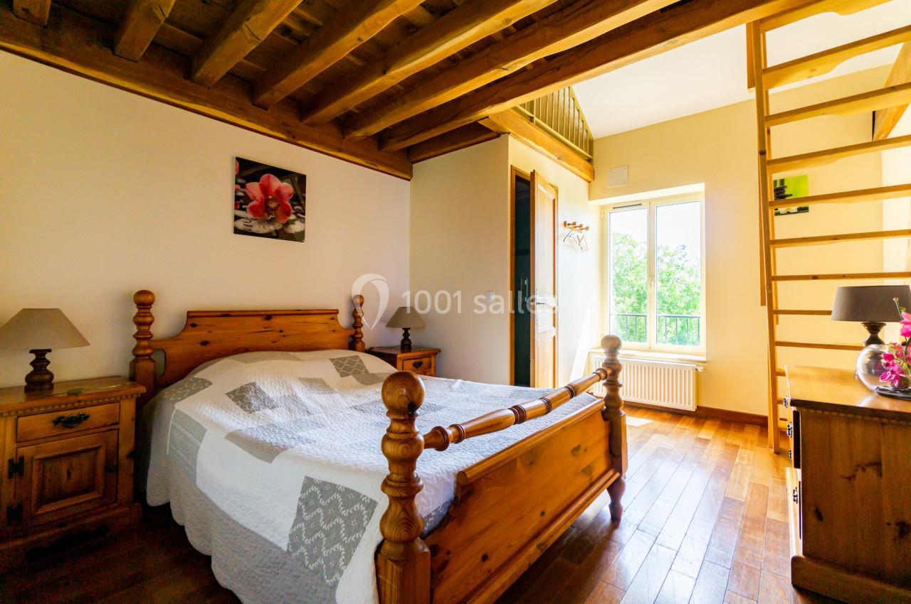 Chambre lumineuse avec lit en bois, plafond en poutres apparentes, échelle menant à une mezzanine et fenêtre donnant sur la…