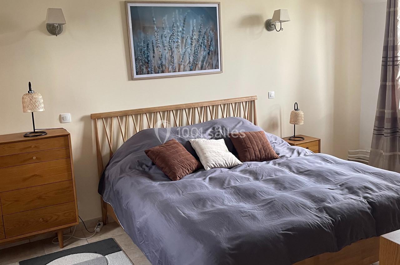 Chambre lumineuse avec lit double, linge gris, coussins décoratifs, mobilier en bois et tableau mural.