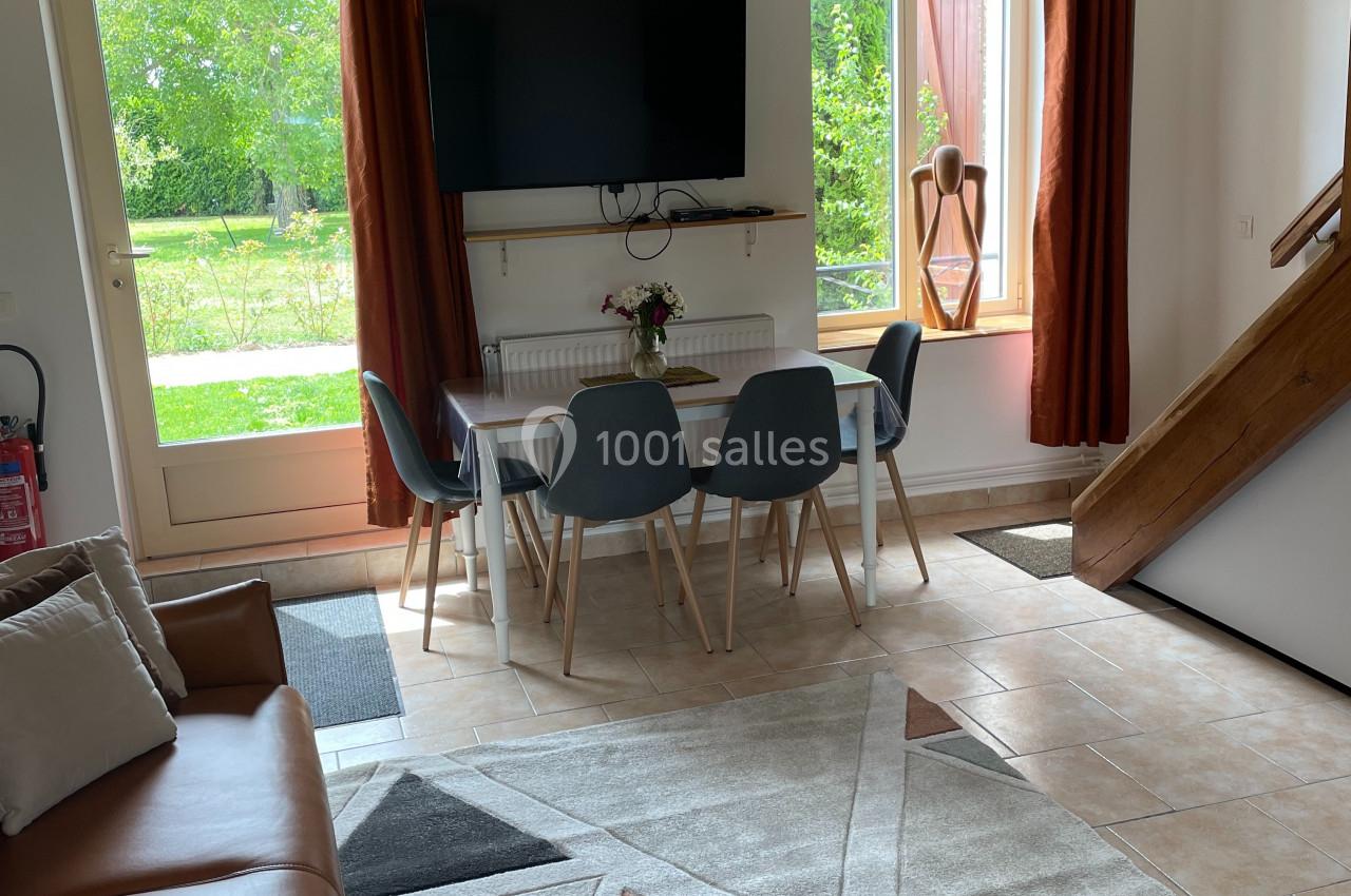 Salon lumineux avec canapé, table à manger, quatre chaises, télévision murale, tapis géométrique et vue sur jardin.