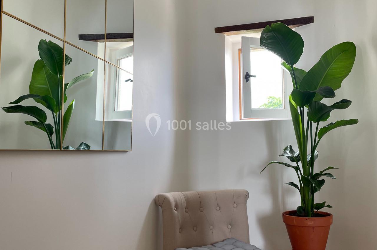 Fauteuil beige avec coussin, plante en pot sur un tabouret et miroir doré dans un coin lumineux avec deux petites fenêtres.