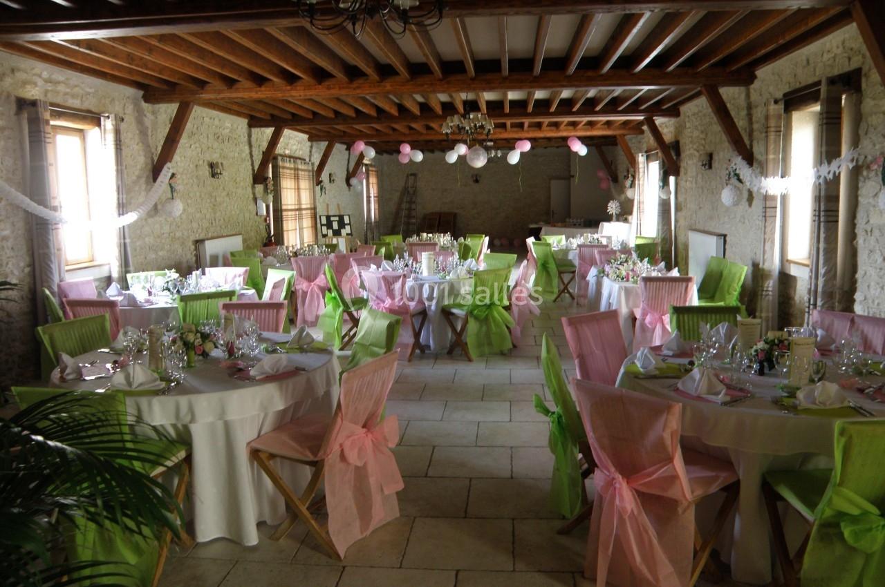 Salle de réception décorée avec des tables rondes, nappes blanches et chaises ornées de nœuds roses et verts.