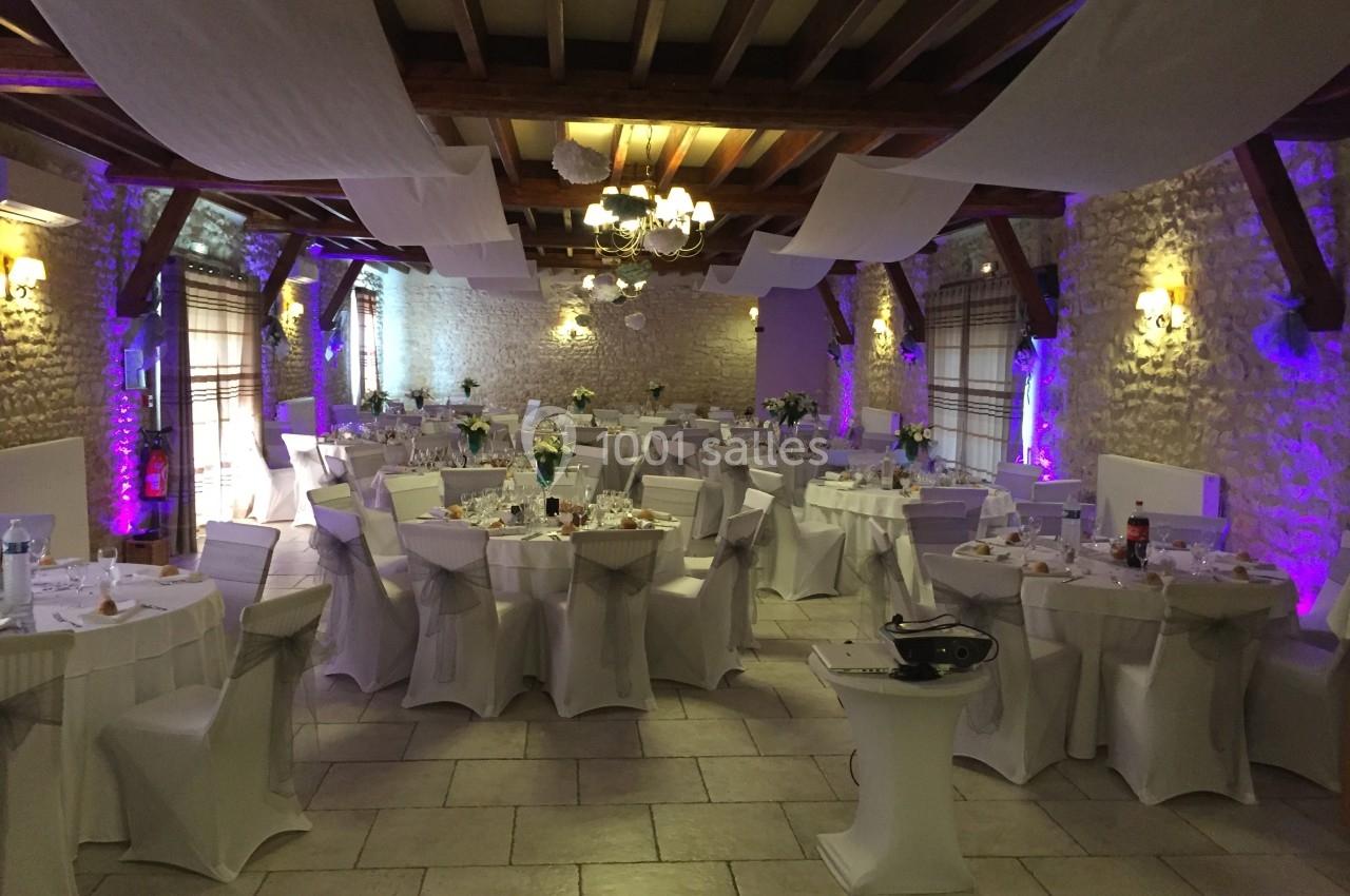 Salle de réception décorée avec des tables rondes nappées de blanc, éclairage tamisé et poutres apparentes.