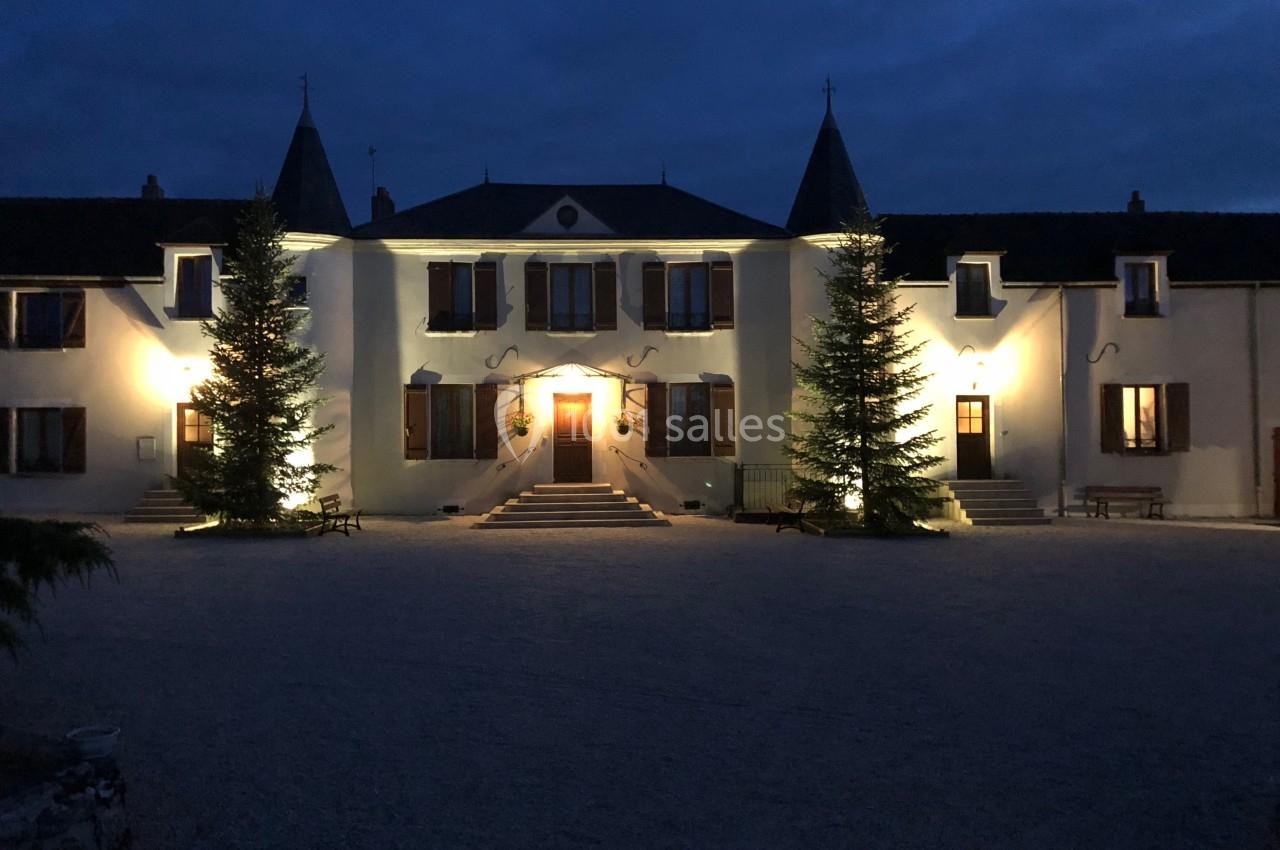 Façade d'un bâtiment éclairé avec des sapins de chaque côté, prise de nuit sous un ciel sombre.