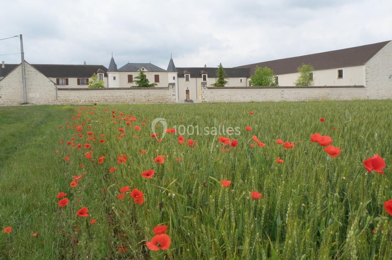 Champ de coquelicots rouges au premier plan avec un bâtiment en pierre et des toits en arrière-plan sous un ciel nuageux.