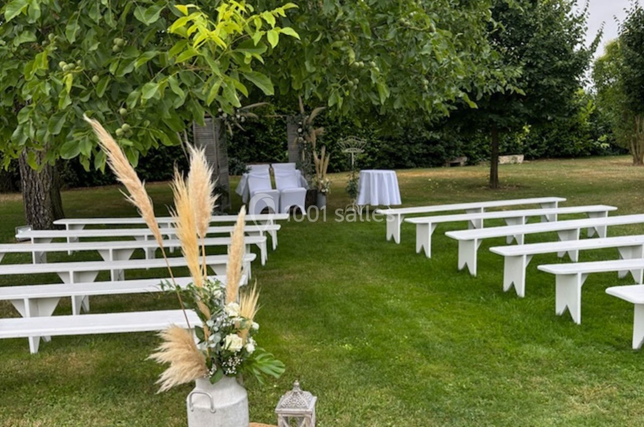 Disposition de bancs blancs en plein air sous des arbres, avec une table décorée pour une cérémonie.