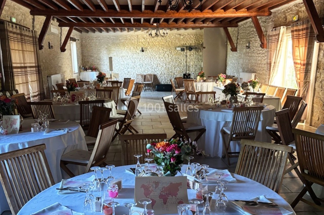 Salle de réception en pierre avec tables rondes décorées de fleurs et chaises en bois, éclairée par la lumière naturelle.