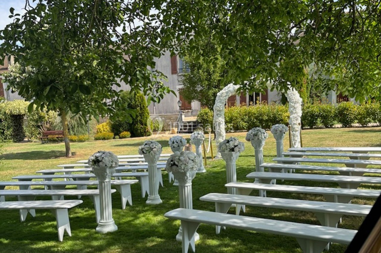 Chaises blanches alignées sous des arbres dans un jardin, décorées de fleurs blanches pour une cérémonie en plein air.