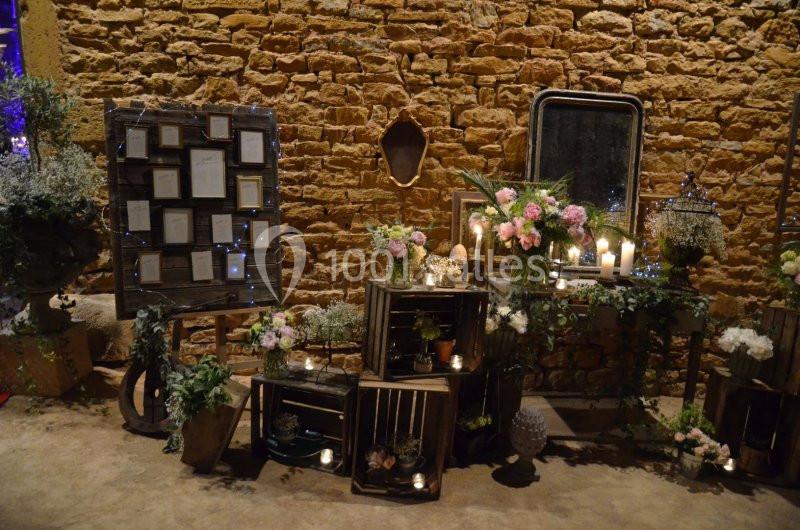 Décoration florale et lumineuse avec des caisses en bois, bougies et cadres, devant un mur en pierre.