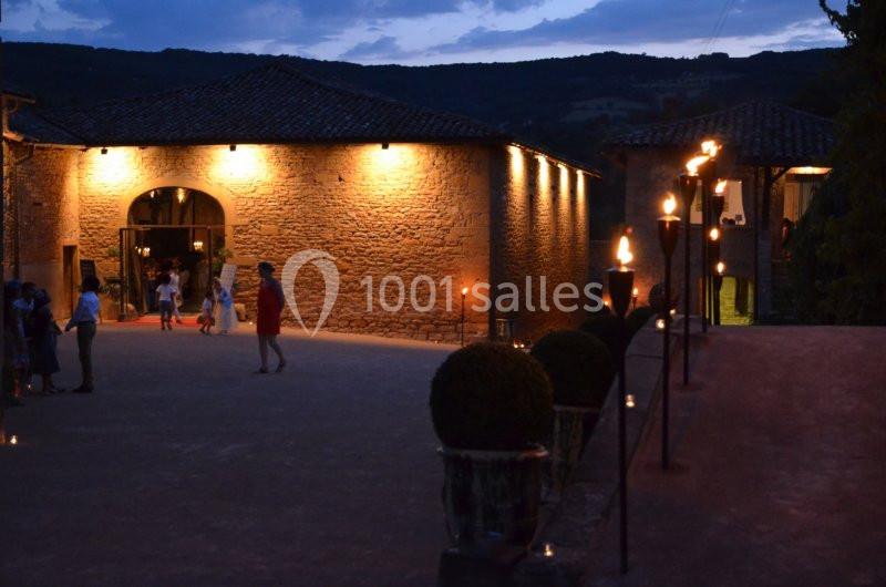 Cour éclairée au crépuscule avec des bâtiments en pierre, des torches allumées et des personnes en arrière-plan.