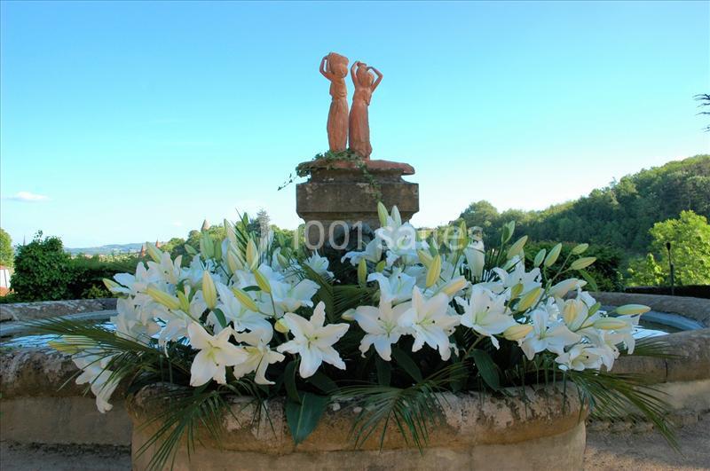 Statue en pierre sur une fontaine entourée de lys blancs et de feuillages, avec un paysage verdoyant en arrière-plan.
