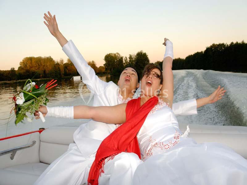 Un couple en tenue de mariage célèbre joyeusement sur un bateau naviguant sur une rivière bordée d'arbres.