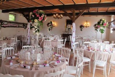 Salle de réception lumineuse avec tables rondes dressées, chaises blanches et décorations florales élégantes.