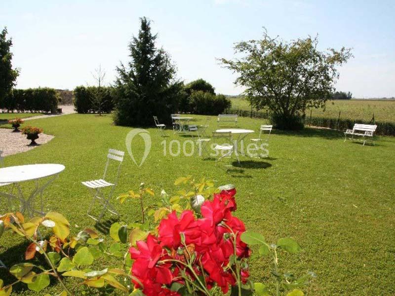 Chaises et tables blanches disposées sur une pelouse avec des fleurs rouges au premier plan et des arbres en arrière-plan.
