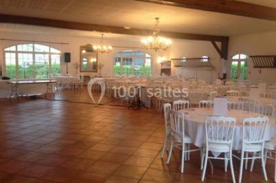 Salle de réception lumineuse avec tables rondes dressées, chaises blanches et décorations florales élégantes.