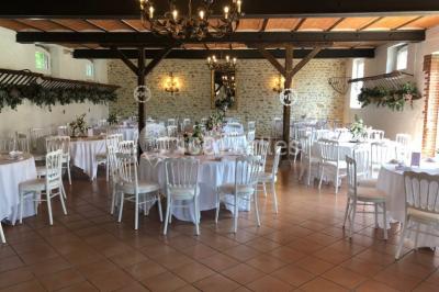 Salle de réception lumineuse avec tables rondes dressées, chaises blanches et décorations florales élégantes.