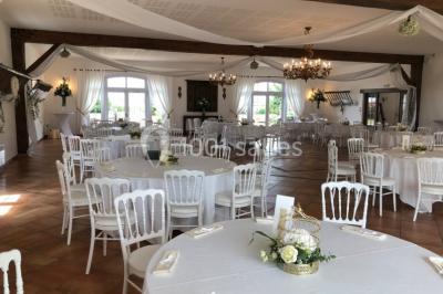 Salle de réception lumineuse avec tables rondes dressées, chaises blanches et décorations florales élégantes.