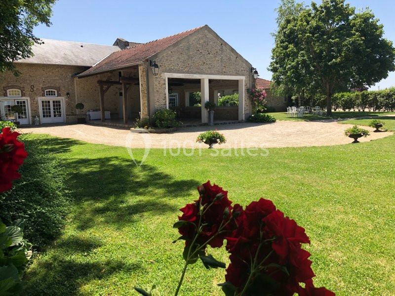 Bâtiment en pierre avec toit en tuiles, entouré de pelouse, fleurs rouges au premier plan et arbres en arrière-plan.